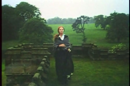Long shot. Entire figure is visible walking through verdant outdoor space.