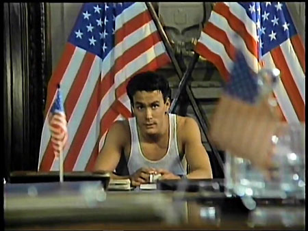 Medium shot. Figure seated at desk with American flags in background.