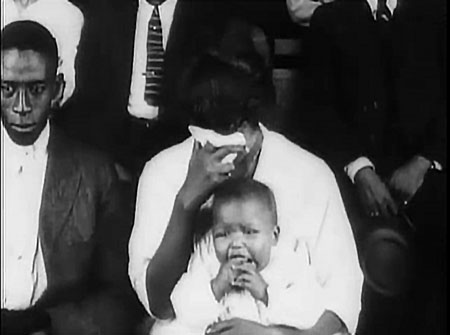 Medium shot of audience members. Main figures are crying baby and woman with head buried in handkerchief.