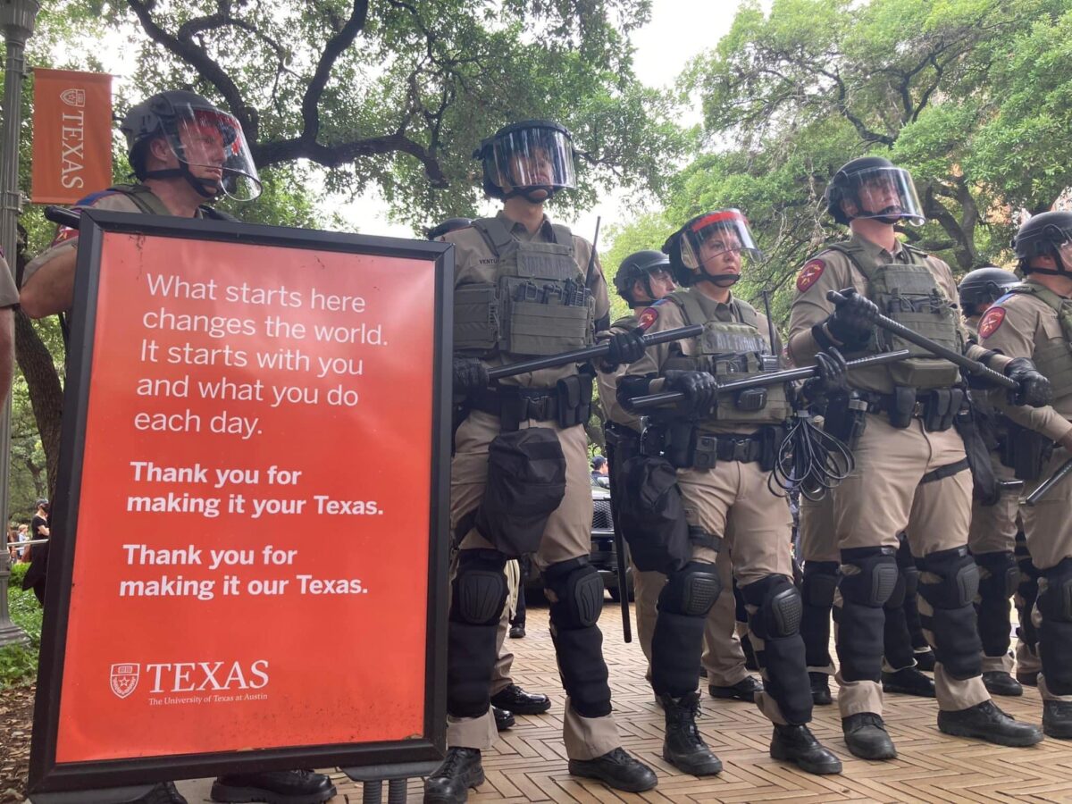 Picture of riot police standing next to a UT Austin sign that says "What starts here changes the world. It starts with you and what you do each day