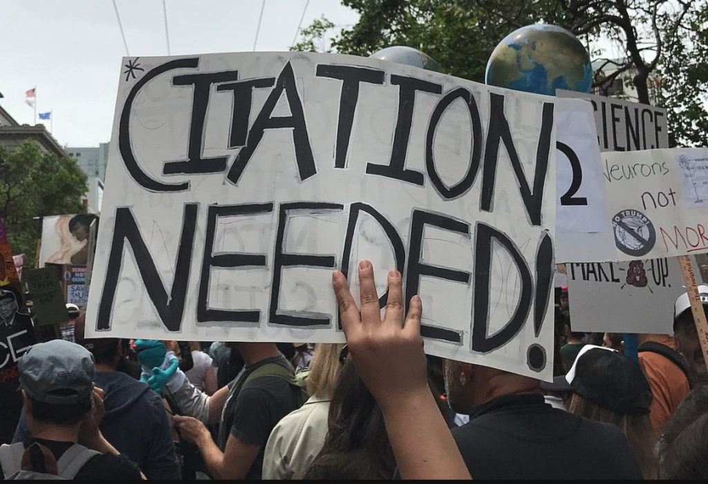 a picture of a protester holding up a sign that reads, "Citation Needed!"